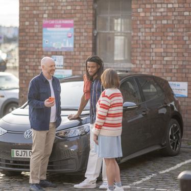 Three people looking at the Zapmap app with an electric car charging behind them