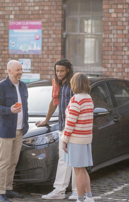 Three people looking at the Zapmap app with an electric car charging behind them